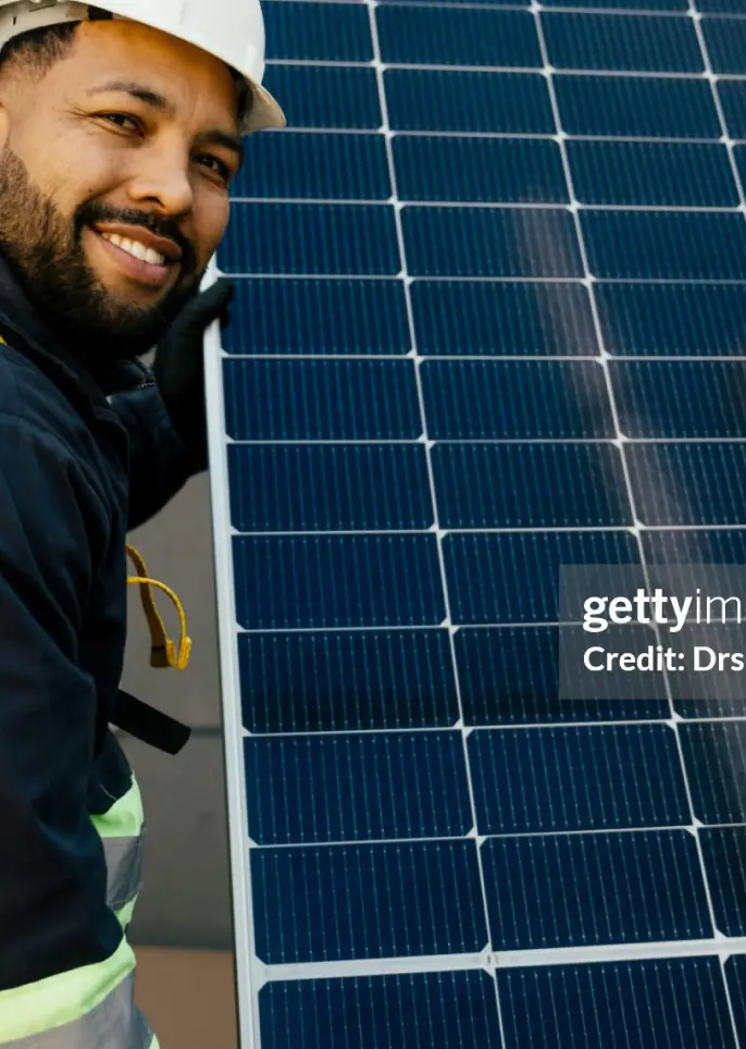 A worker wearing safety gear holds a solar panel