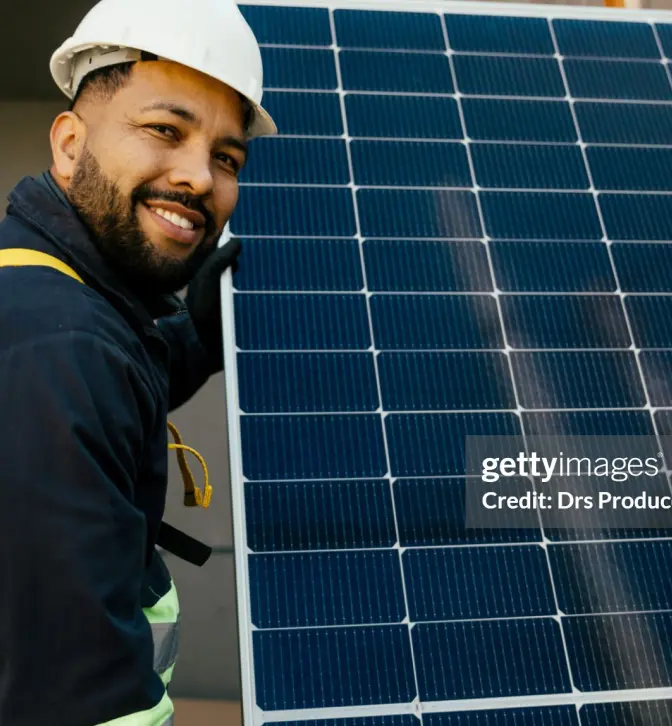 A worker wearing safety gear holds a solar panel